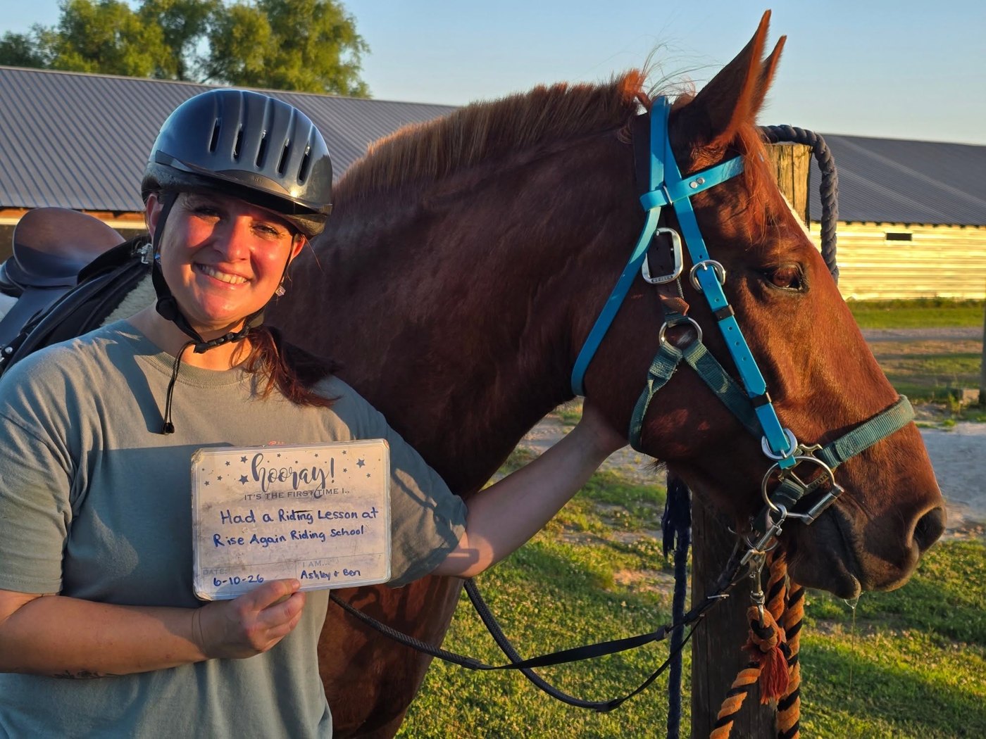 A rider beaming beside her horse, holding a card that reads 'Hooray, it's the first time I had a riding lesson at Rise Again Riding School.'
